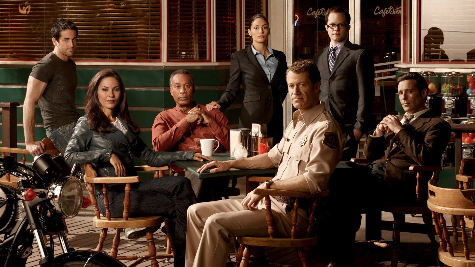 Group of seven adults in a retro diner, some seated at a table and others standing, with a motorcycle visible in the foreground.