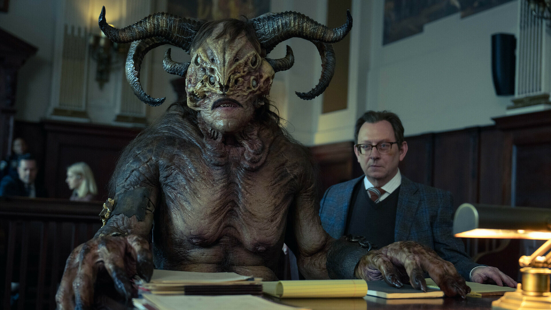evil-3 | film.org.pl Horned, horned demon-like creature sits at a judge’s bench-style table in a courtroom, with a suited man nearby and court paper stacks in foreground.