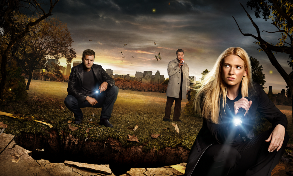 Three adults in a park at dusk investigate a large cracked hole in the ground; a blonde woman on the right points a flashlight, a man in a leather jacket crouches left with a light, and another man in a trench coat stands in the middle.