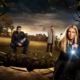 Three adults in a park at dusk investigate a large cracked hole in the ground; a blonde woman on the right points a flashlight, a man in a leather jacket crouches left with a light, and another man in a trench coat stands in the middle.