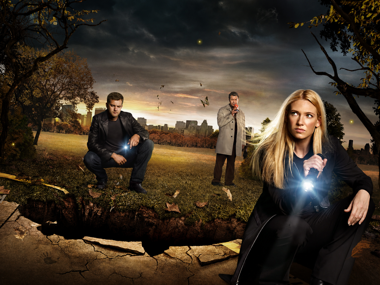 Three adults in a park at dusk investigate a large cracked hole in the ground; a blonde woman on the right points a flashlight, a man in a leather jacket crouches left with a light, and another man in a trench coat stands in the middle.