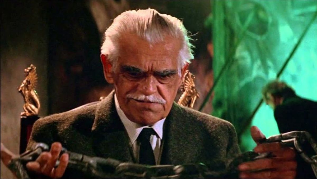 Older man in a brown blazer and tie, with white hair and mustache, examining snakes wrapped around his hands in a dim setting.
