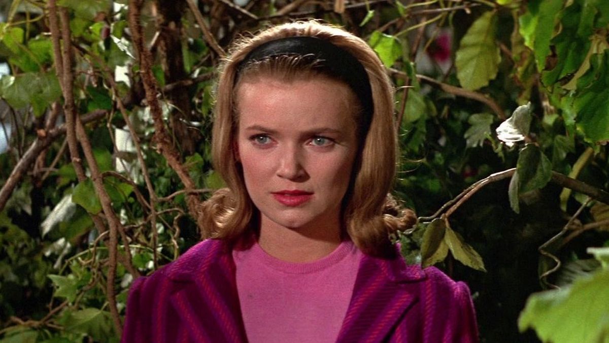 Woman with blonde hair wearing a pink top and purple jacket, standing among green vines and leaves.