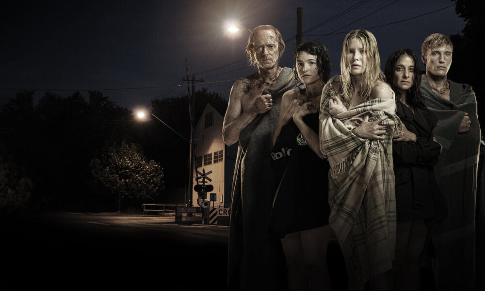 Five people stand close together at night, wrapped in blankets, on a dark street with a dim streetlight in the background, looking toward the camera.