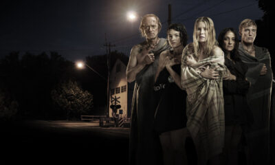 Five people stand close together at night, wrapped in blankets, on a dark street with a dim streetlight in the background, looking toward the camera.