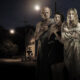 Five people stand close together at night, wrapped in blankets, on a dark street with a dim streetlight in the background, looking toward the camera.