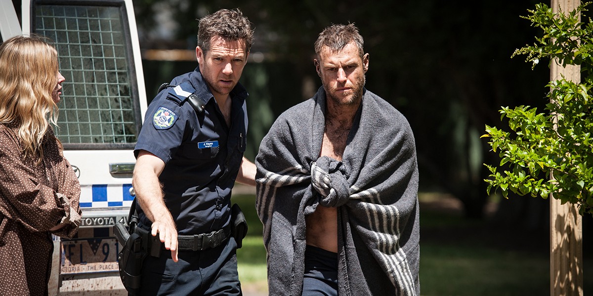 Officer leads a shirtless man wrapped in a blanket away from a police vehicle while a woman watches nearby.