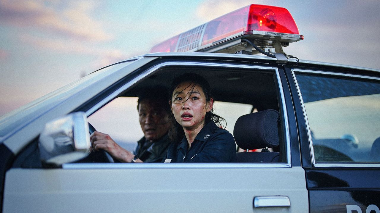 hope-3 | film.org.pl Female police officer in the passenger seat looks surprised, while a male officer drives; red police light on the roof glows, during sunset.