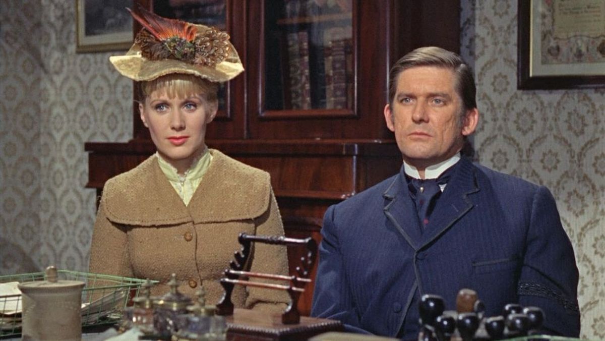 Woman wearing a vintage hat and a tan coat sits next to a stern-looking man in a blue suit at a wooden desk in a retro-style room.