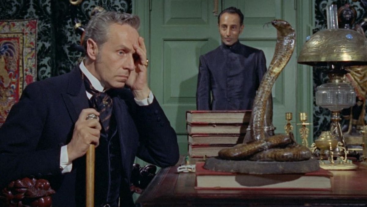 A man in a dark suit looks distressed while resting a hand on his temple, holding a cane, in a room with a large snake sculpture on the desk and a standing figure in the background.