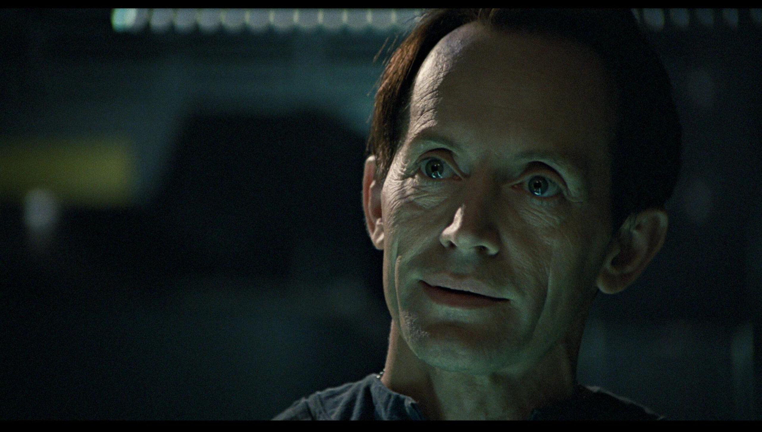 Close-up of a man in dim lighting, eyes wide with concern, looking upward/forward with a tense expression.