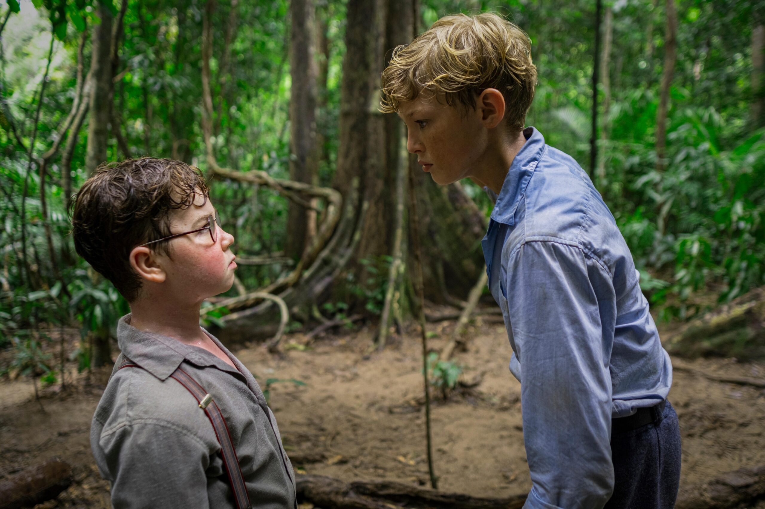 Two boys in a jungle face each other, one with glasses looking up at the taller boy in a blue shirt during a tense conversation in a dense forest.