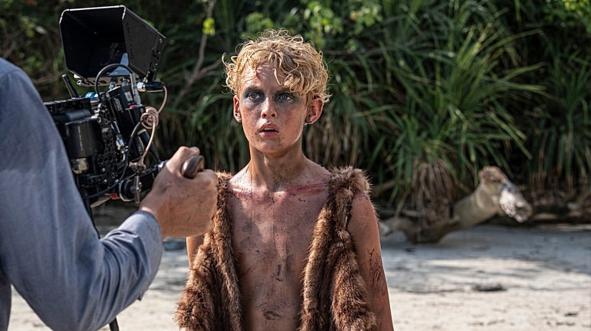 Actor with dirt on chest and face, wearing a brown fur vest, stands on a sandy beach as a camera operator films him on a movie set.