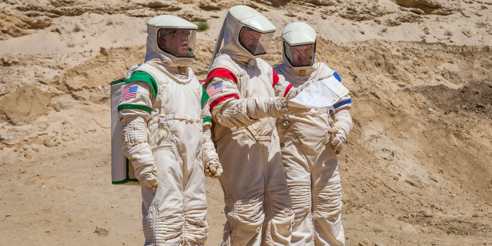 moonbase-8 | film.org.pl Three astronauts in beige spacesuits with U.S. flag patches study a map in a rocky desert setting.