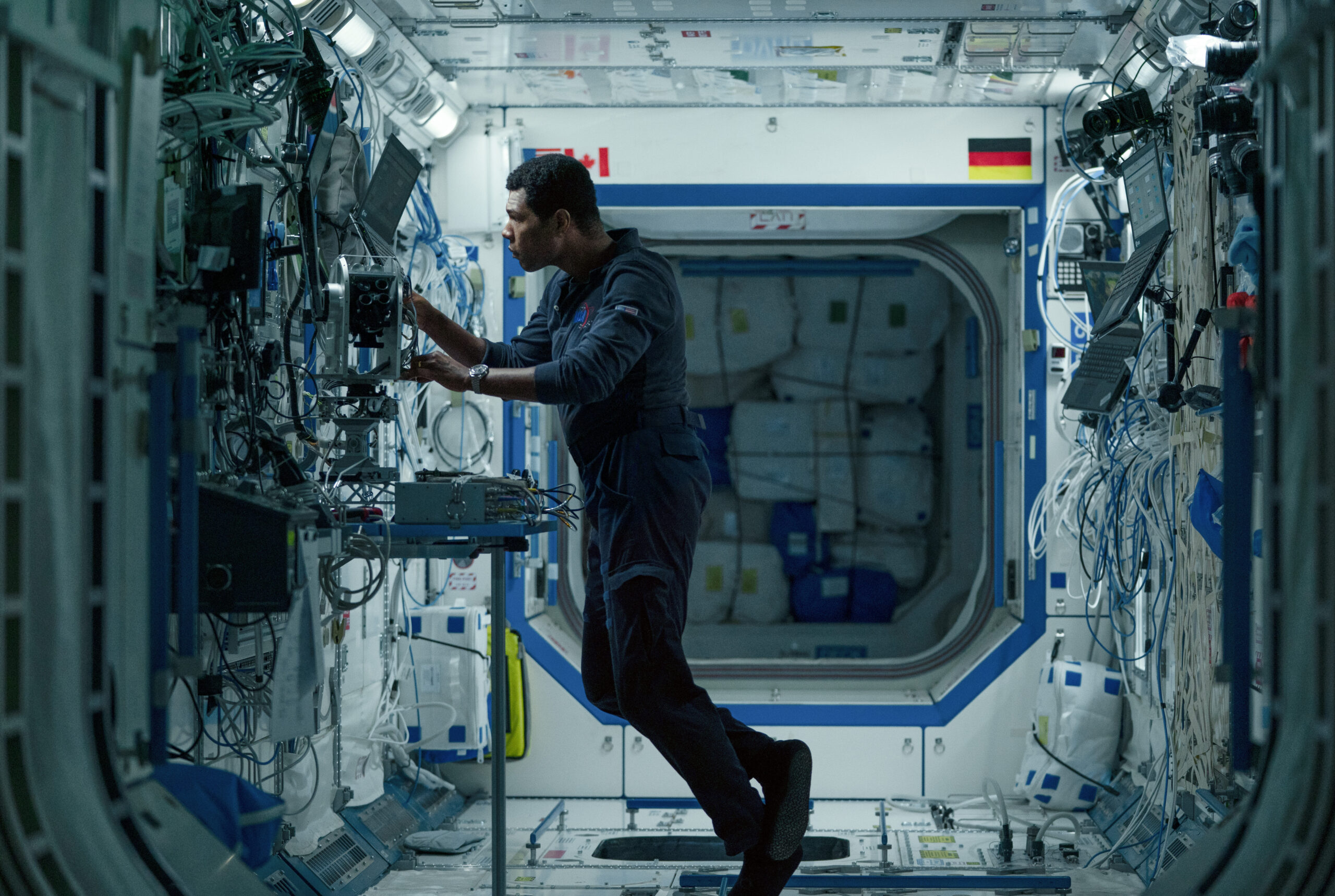 Engineer in a blue jumpsuit adjusting wiring and equipment inside a crowded space station corridor full of cables and control panels.