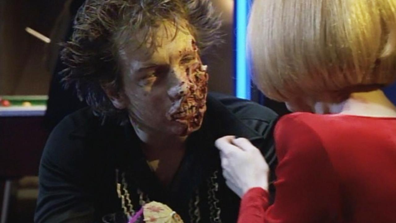 Man with half-face zombie makeup sits facing a woman in red who touches his shoulder in a dim setting.
