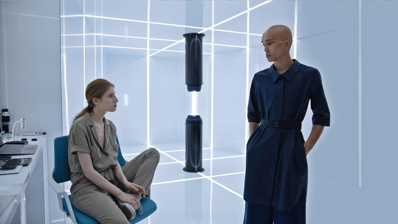 osmosis-2 | film.org.pl A woman in a beige jumpsuit sits on a blue chair and talks with a bald man in a dark blue outfit in a futuristic, neon-lit lab with a tall cylindrical device between them.