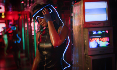Person in a black leather outfit with a glowing blue visor, standing in a neon-lit nightclub environment.