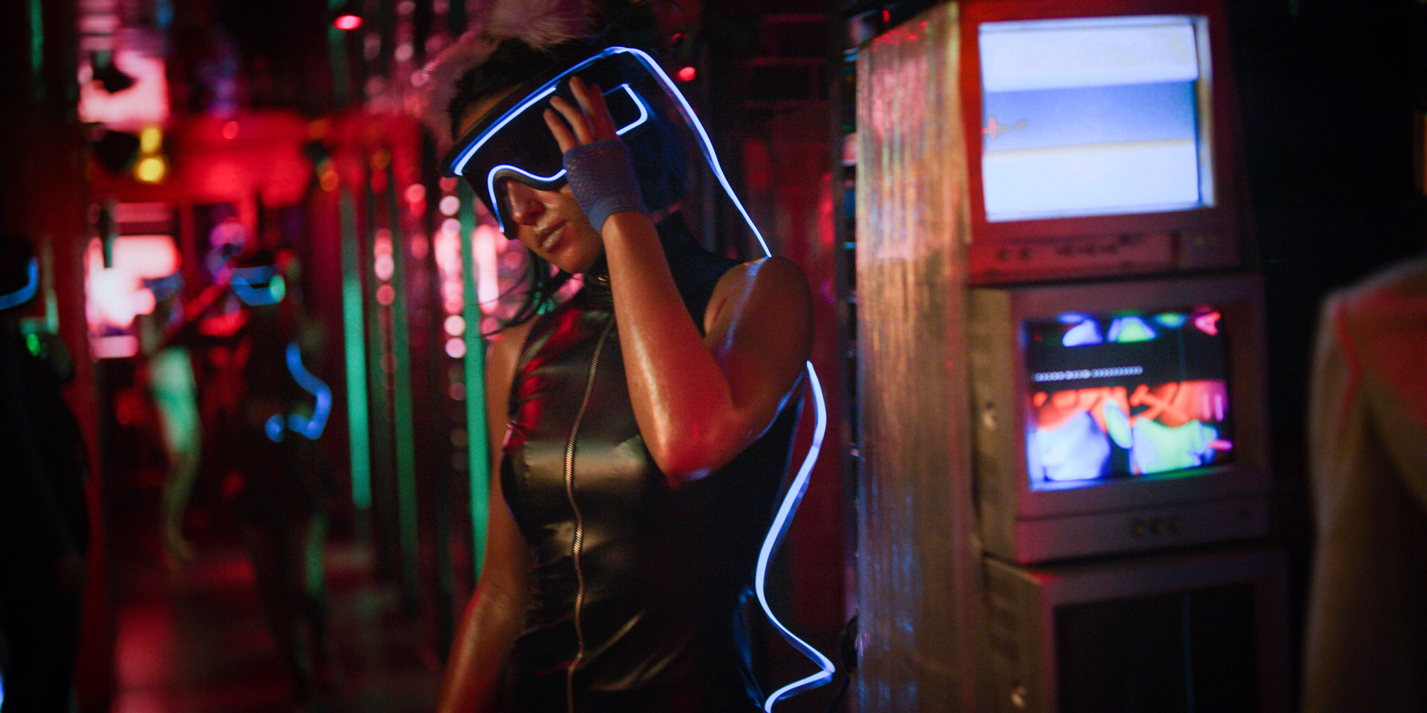 Person in a black leather outfit with a glowing blue visor, standing in a neon-lit nightclub environment.