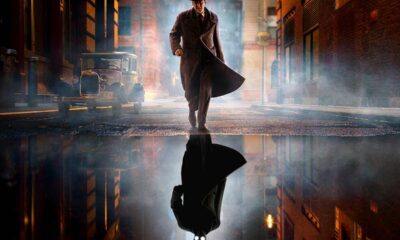 Noir-styled man in a long trench coat and fedora walks down a foggy city street at night, his reflection visible in a puddle.