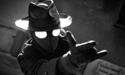 Monochrome image of a figure in a fedora and trench coat wearing a gas mask with bright eye lenses, reaching forward with outstretched arms patrolling toward the viewer.