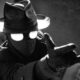 Monochrome image of a figure in a fedora and trench coat wearing a gas mask with bright eye lenses, reaching forward with outstretched arms patrolling toward the viewer.