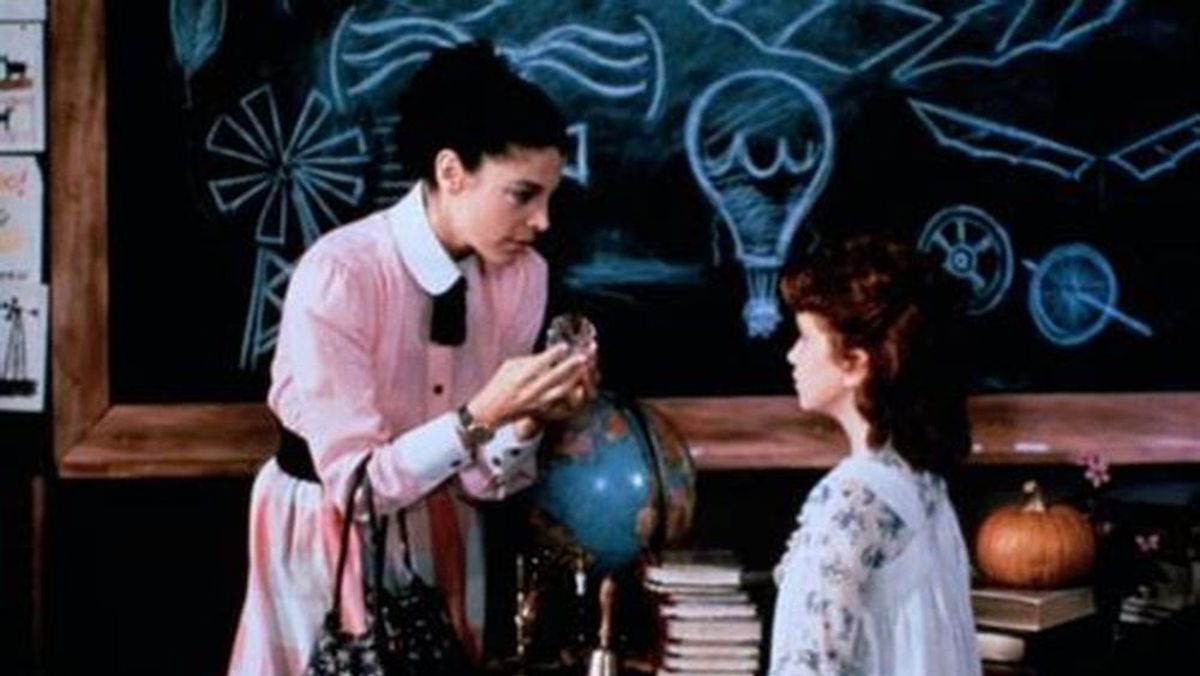 A teacher in a pink dress shows a small object to a young girl in a classroom, with a globe, stacked books, and chalkboard drawings in the background.