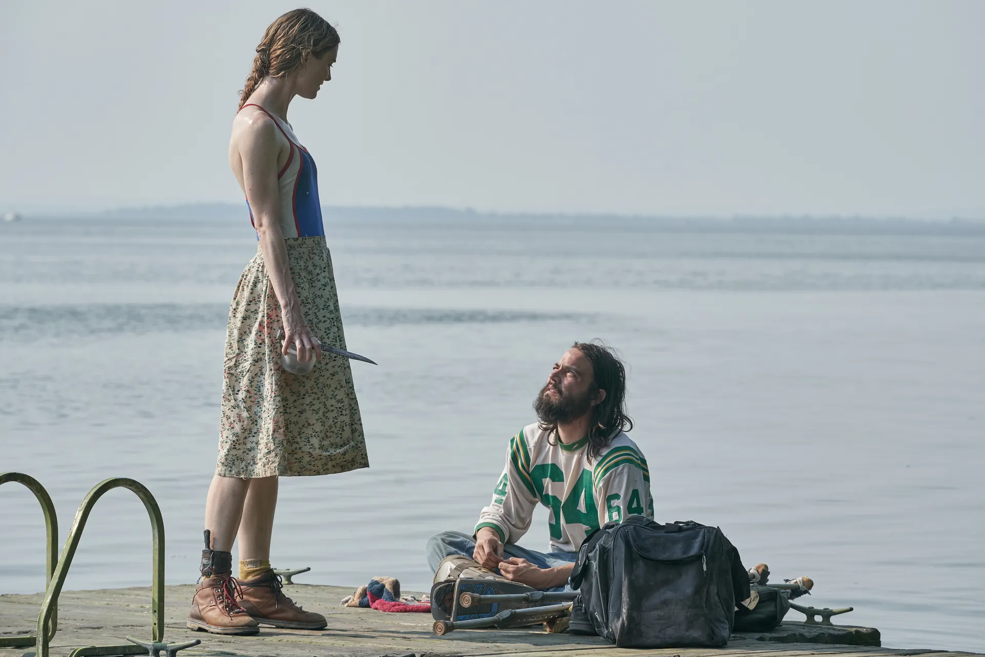 Tense scene on a dock where a woman in a floral skirt and boots stands over a kneeling bearded man holding a knife pointed at him by the water edge.