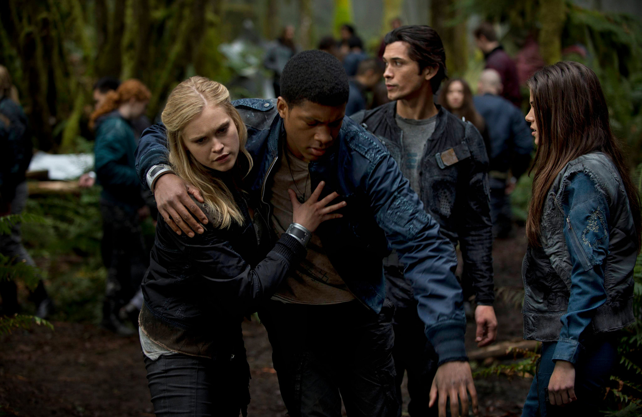 A group of young adults in a forested area supports a friend who appears distressed, with others standing nearby in jackets.