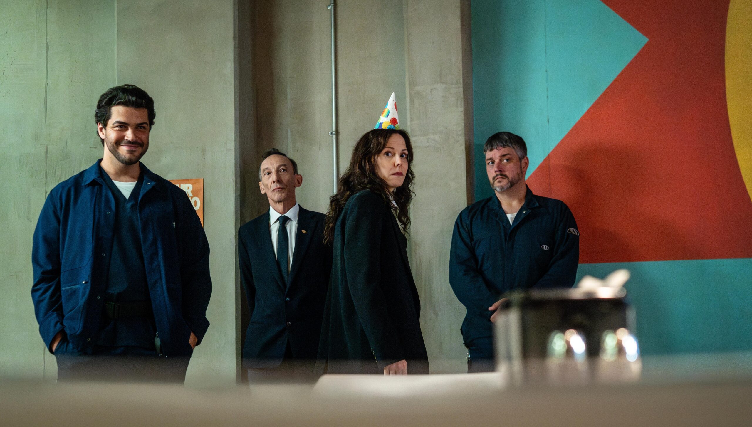Four adults in dark blue uniforms stand indoors against a concrete wall; a woman wearing a colorful party cone hat looks toward the camera.