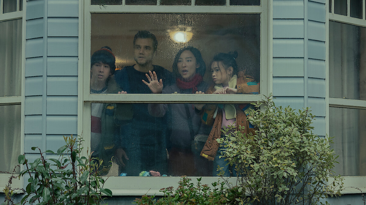 Four people lean against a large bay window, peering outside with curious or worried expressions.