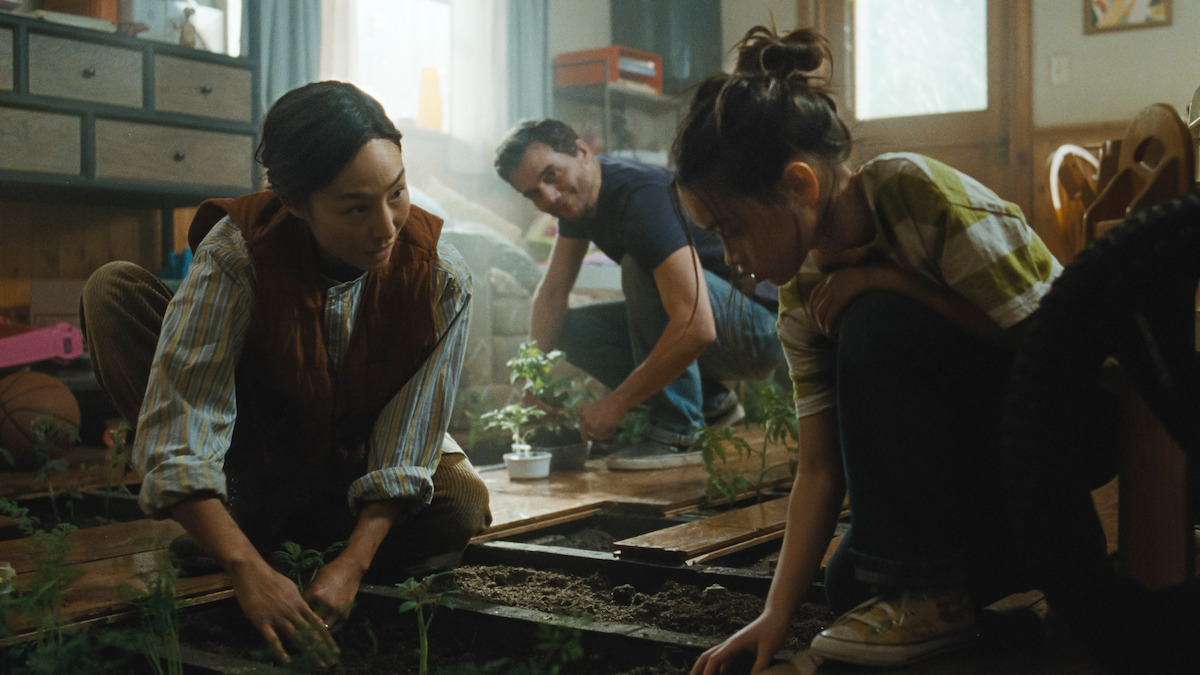 Three people kneeling by wooden planters, tending young plants in a sunlit indoor garden room, casual and collaborative scene.
