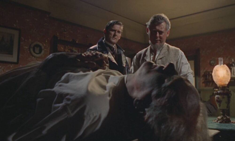 Two men stand beside a bed where a person lies unconscious in a dim, vintage bedroom scene, looking concerned (movie still).