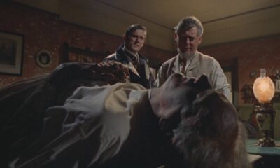 Two men stand beside a bed where a person lies unconscious in a dim, vintage bedroom scene, looking concerned (movie still).