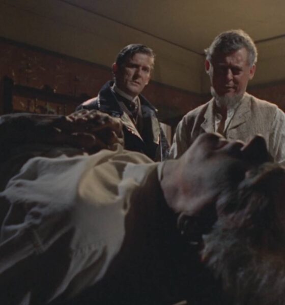 Two men stand beside a bed where a person lies unconscious in a dim, vintage bedroom scene, looking concerned (movie still).