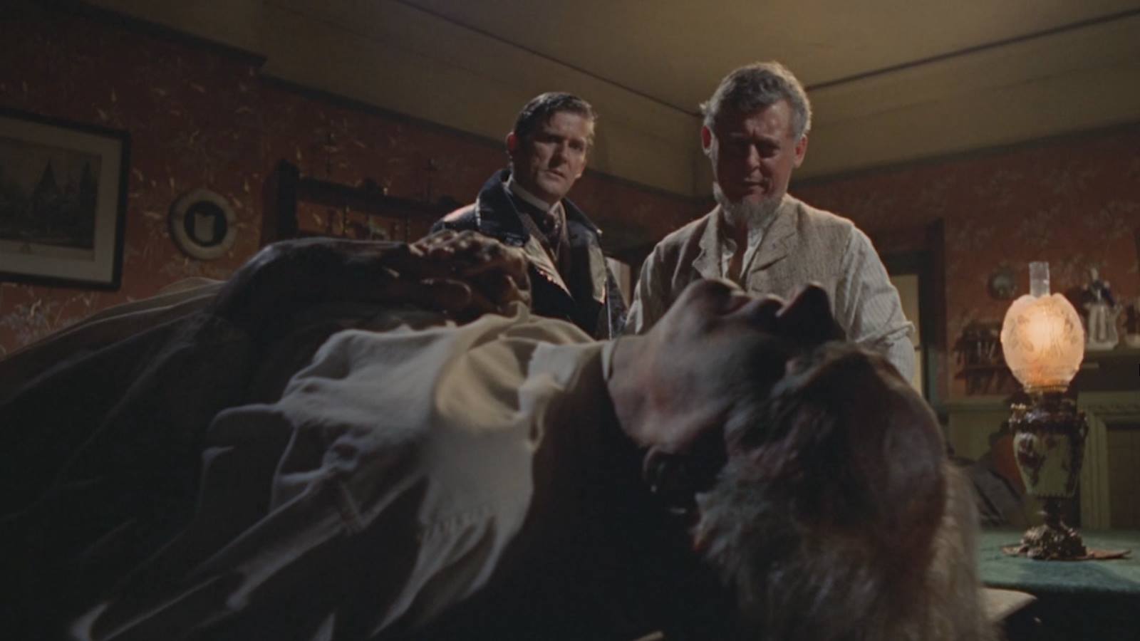 Two men stand beside a bed where a person lies unconscious in a dim, vintage bedroom scene, looking concerned (movie still).