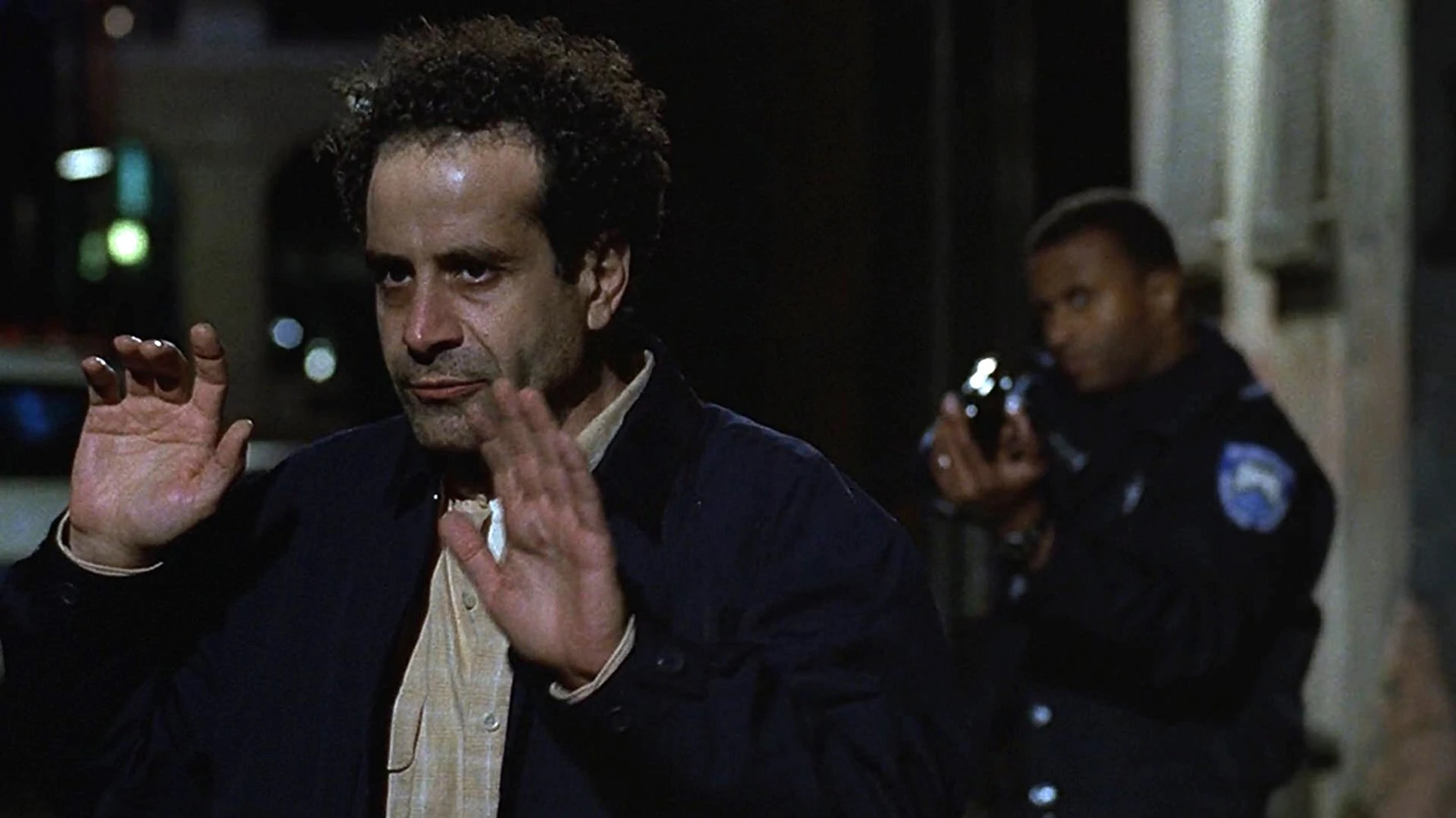 Man in a dark jacket raises both hands in front of him at night, while a police officer with a camera stands behind him in a dim alley.
