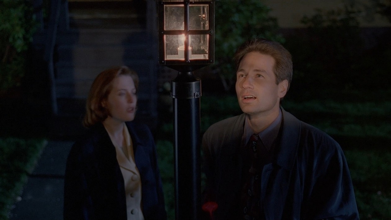 Man in a suit looks up at a lit lantern while a woman in a beige coat stands beside him at night outside.