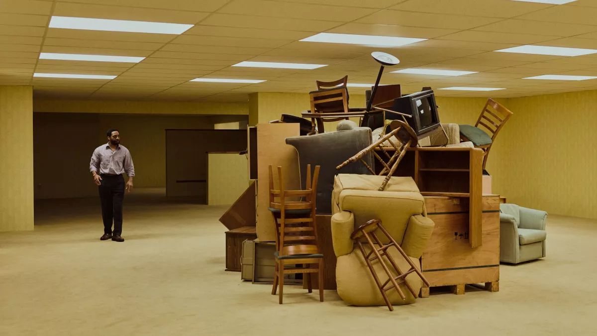 Backrooms 1 | film.org.pl A man stands apart from a massive pile of stacked furniture—chairs, sofas, and cabinets—in a bare yellow-lit room.