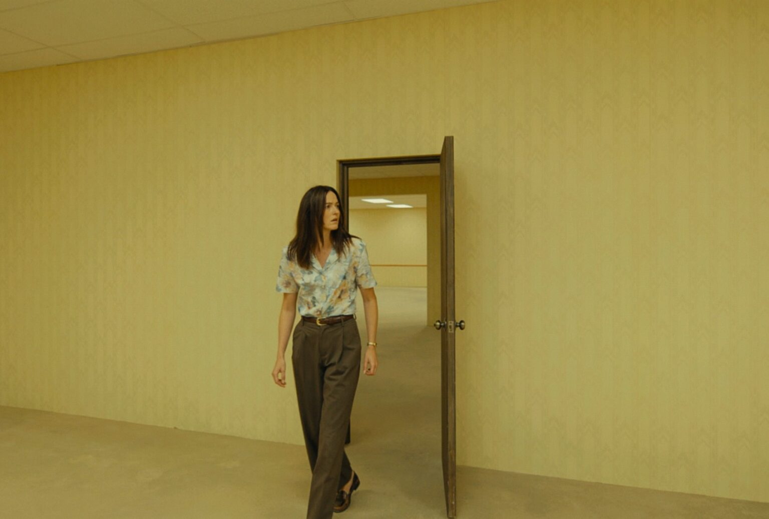Backrooms 2 | film.org.pl Woman in a floral shirt and brown pants walks through an open wooden door into a pale yellow hallway.
