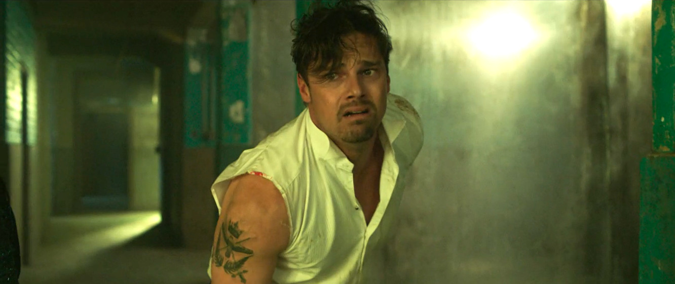 Man in a white shirt looks distressed as smoke fills a dim hallway, reaching forward instinctively.