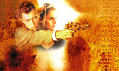 Man in a beige jacket aims a pistol with a bright muzzle flash, orange fiery background fading around him with a woman behind him.