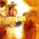 Man in a beige jacket aims a pistol with a bright muzzle flash, orange fiery background fading around him with a woman behind him.
