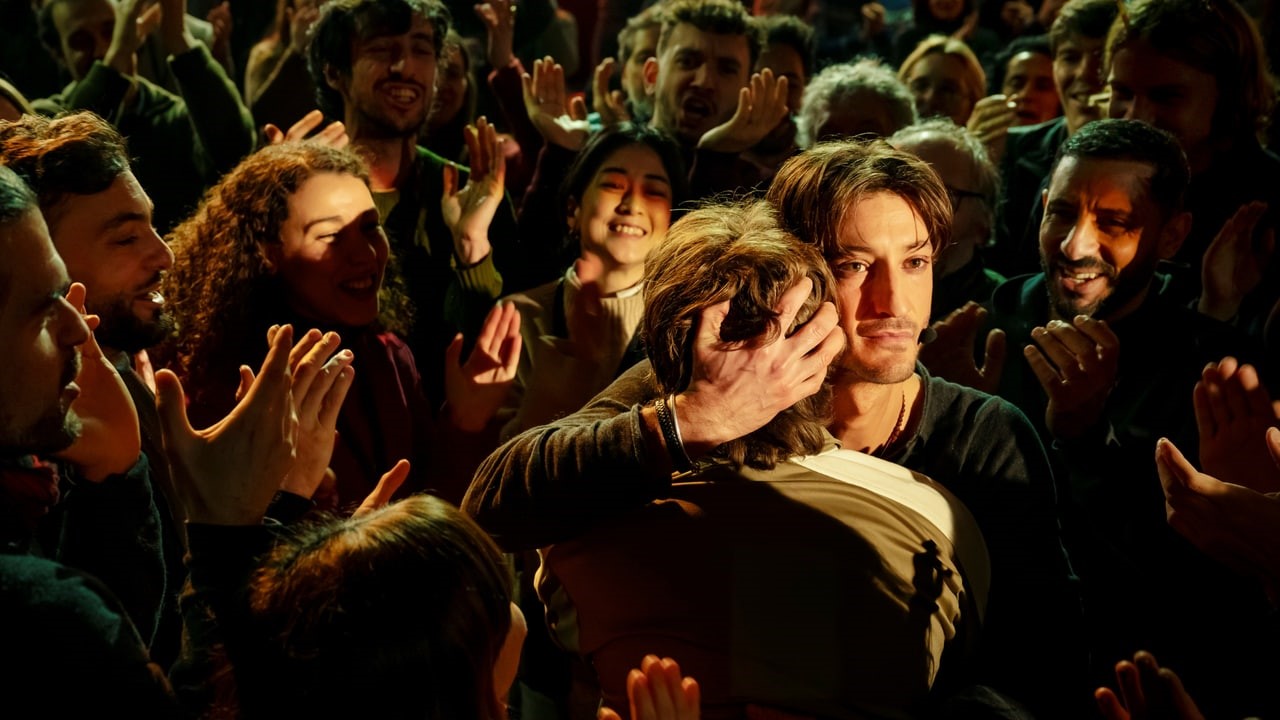 A man hugs a person in the center while a cheering crowd surrounds them at a lively event of applause and smiles.
