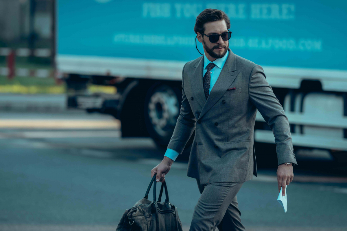 Man in a gray suit and sunglasses walks with a duffel bag beside a blue truck, holding a mask in one hand.