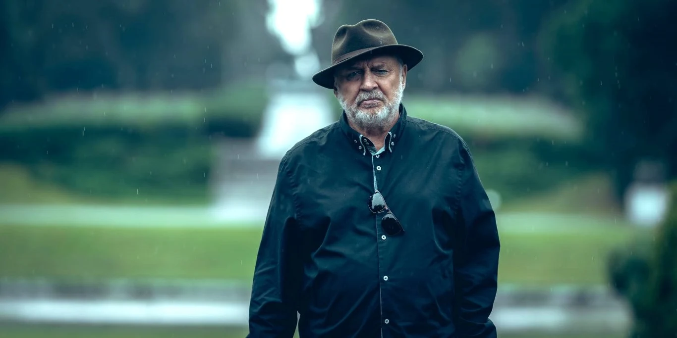 A somber older man in a dark button-down shirt and fedora stands in light rain outdoors, looking ahead.