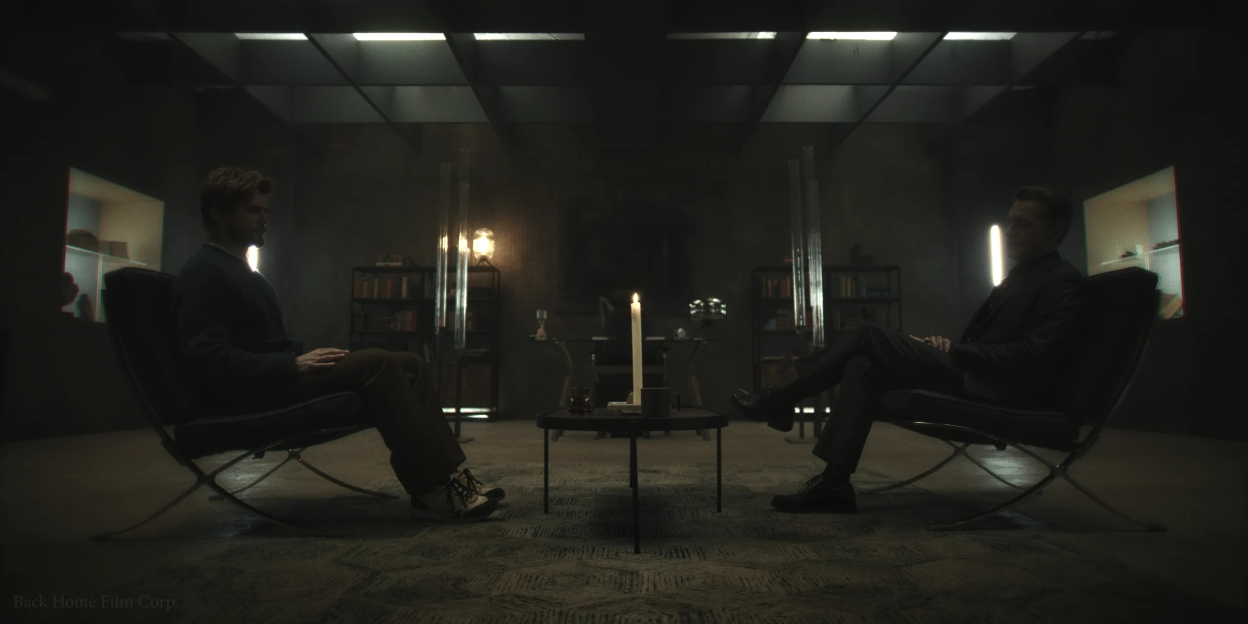 Two men sit in dark, moody room facing each other in modern lounge chairs; a single candle on the table between them creates atmosphere.