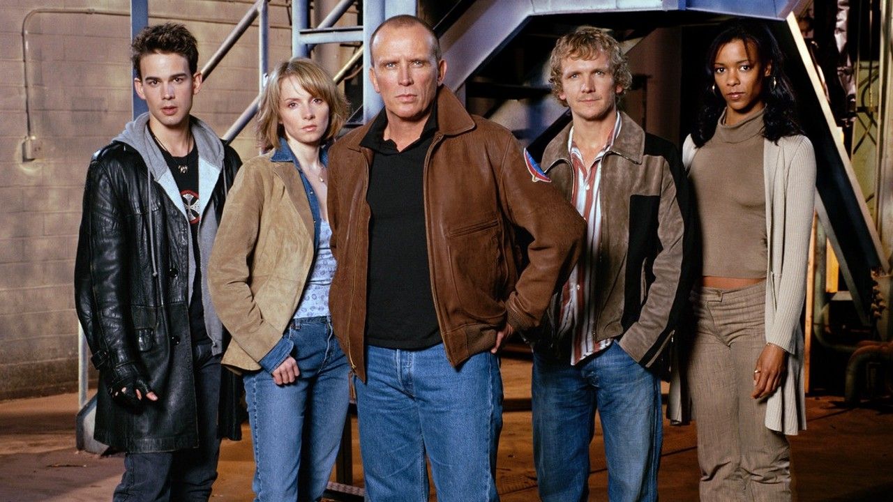 Six adults stand together in an industrial setting, posing for a group photo, wearing casual jackets and jeans.