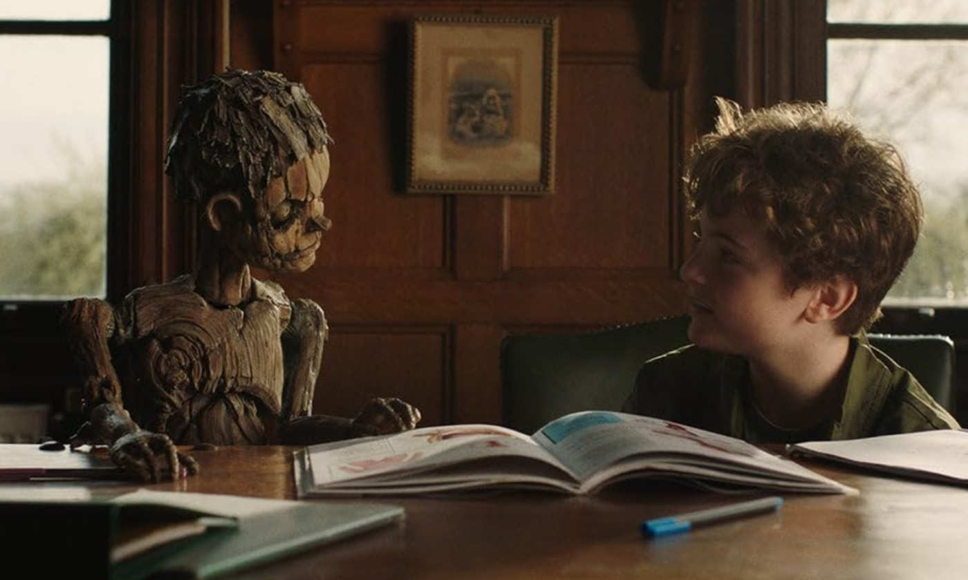 Young boy sits at a wooden desk, reading an open book beside a carved wooden mummy sculpture in a sunlit room.