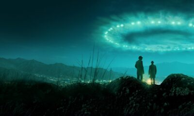 Two men in long coats and fedora hats stand on a rocky hill, looking toward a valley city and distant mountains under a teal, glowing sky ring.